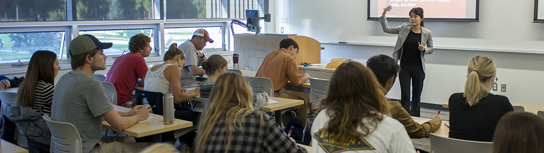 A professor lectures in front of a class, her slides projected behind her.