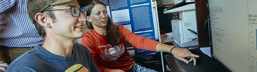 Two engineering students discuss data on a computer screen, a research poster in the background.