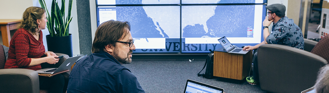 Students and a professor overview a map on a large digital screen.