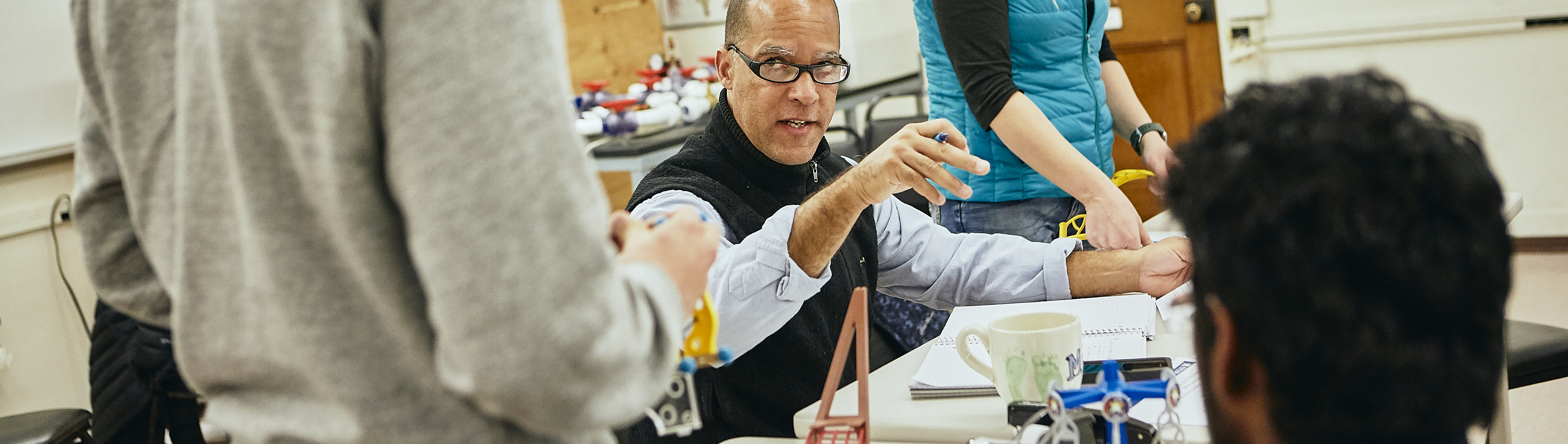 A middle-aged man in a vest and rolled-up shirtsleeves lectures in a graduate Industrial Engineering lab while students look on.