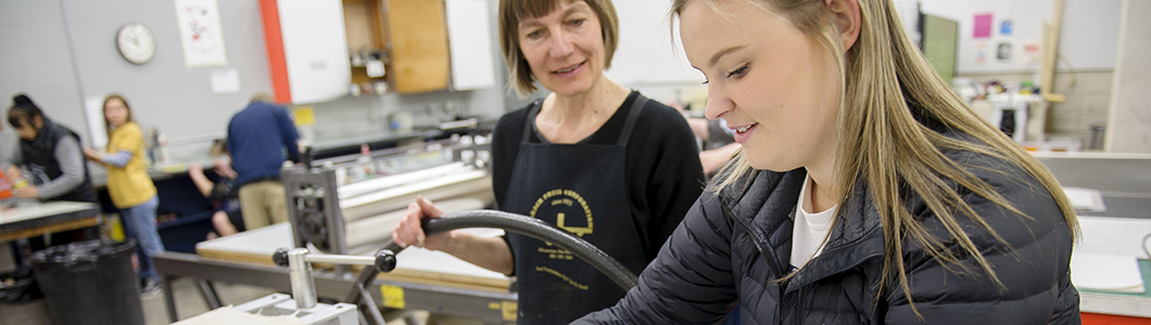 A student oversees funneling a print through a printing press while a professor looks on.