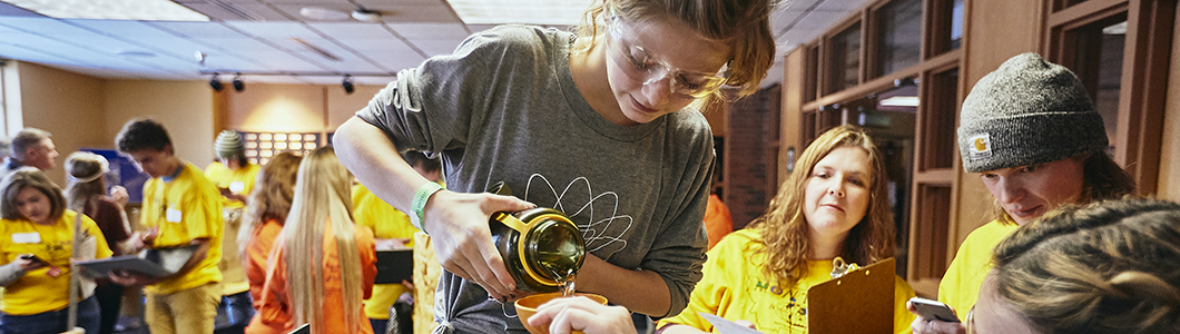 A young student pours liquid into a vessel while a science teacher looks on.