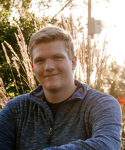 Young male with short golden brown hair and a blue long sleeve shirt. Grasses, trees and birdhouse behind.