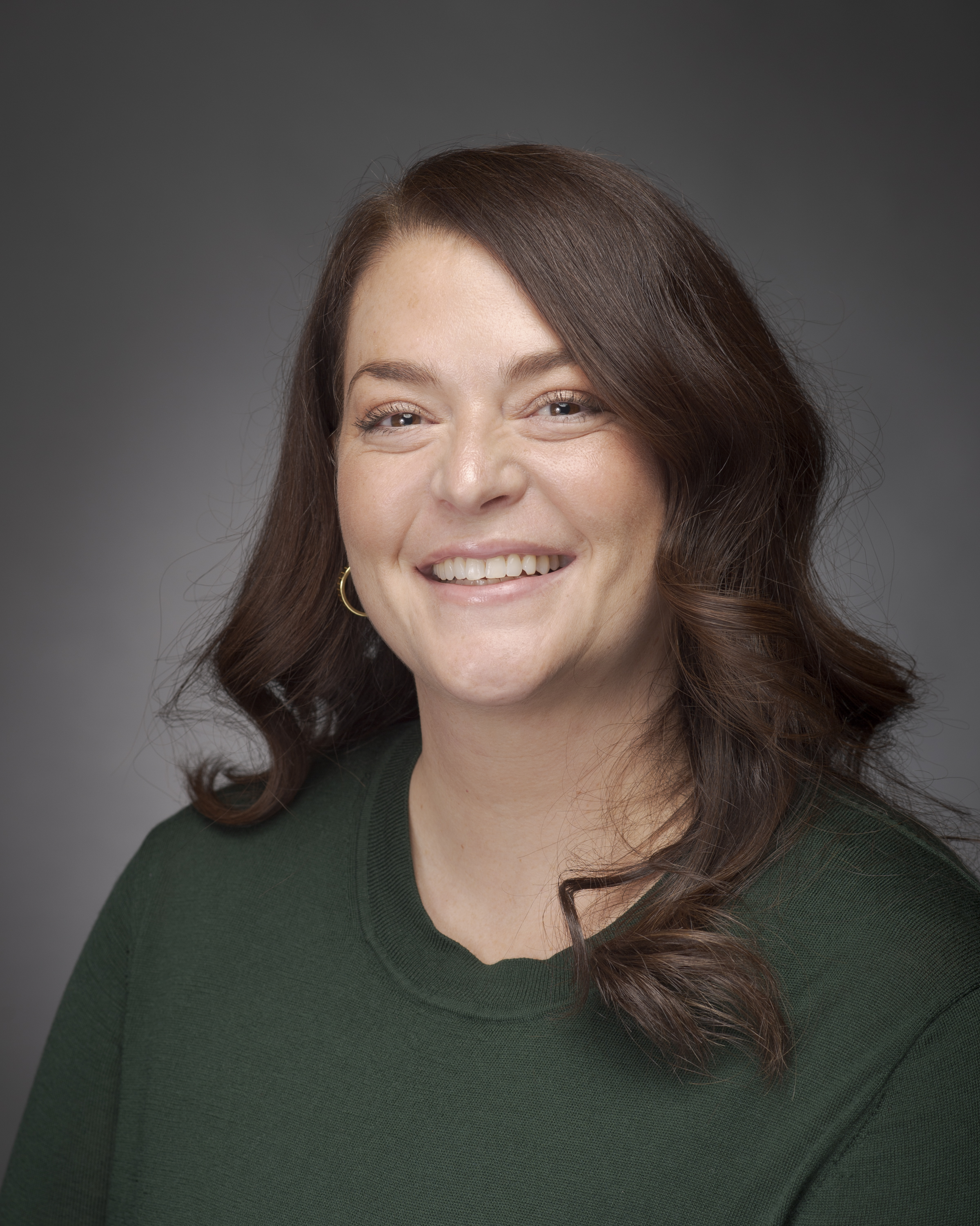 Photo of woman with white skin, brown hair, and a green shirt. The background is gray.