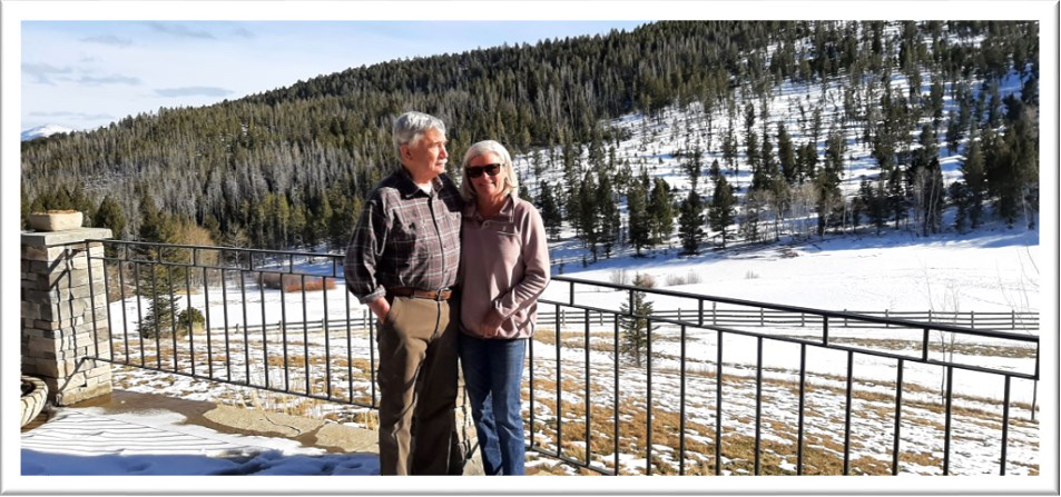 A man and a woman stand together on a stone patio overlooking a vast, snowy mountain valley
