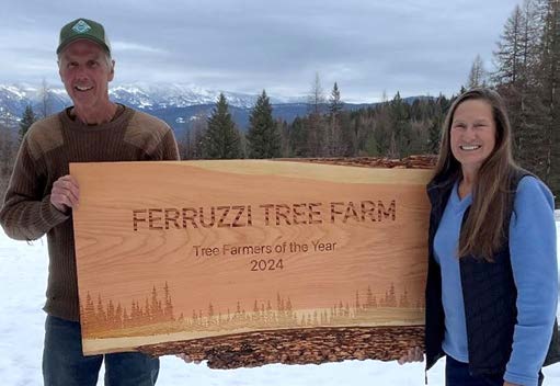 Ferruzzi Tree Farm a photograph of a man and woman standing outside in the snow with trees and mountains in the background. They are holding a large wooden sign that reads, Ferruzzi Tree Farm Tree Farmers of the Year 2024.