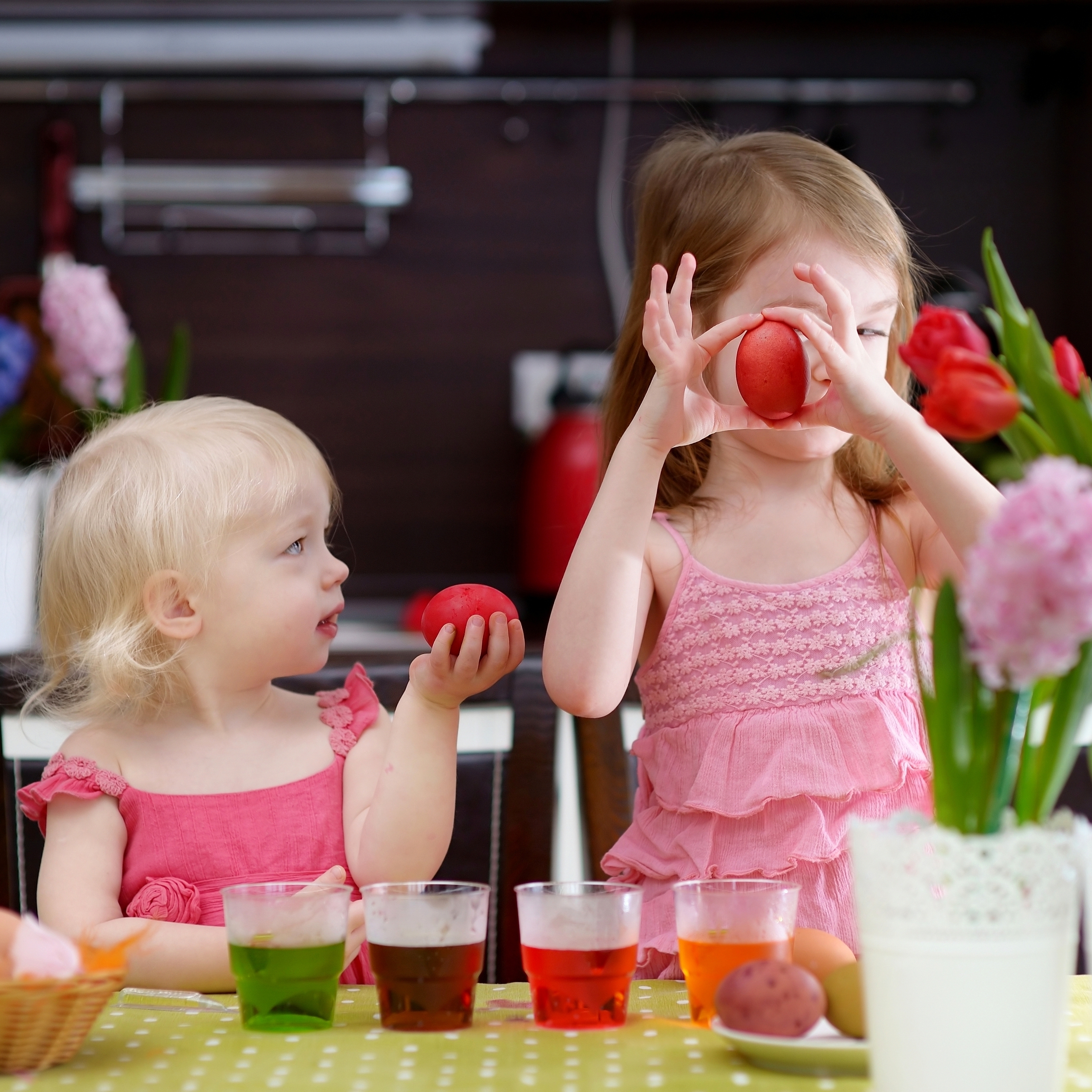 Two children holding red-dyed Easter eggs while sitting at a table with four cups of Easter egg dye.