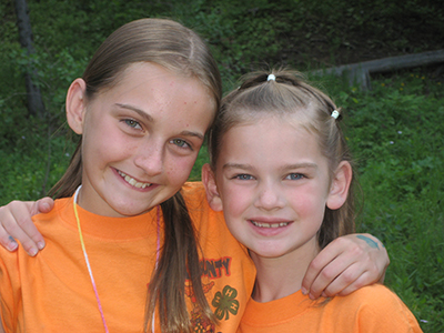 Two children wearing matching T-shirts stand outdoors with their arms around each other. Trees, grass, and a wooden fence are visible in the background.