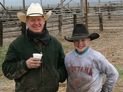 Passing the Torch feature photo An older man and a boy stand side by side in front of a wooden corral and smile at the camera. Both are wearing cowboy hats and are covered in dirt.