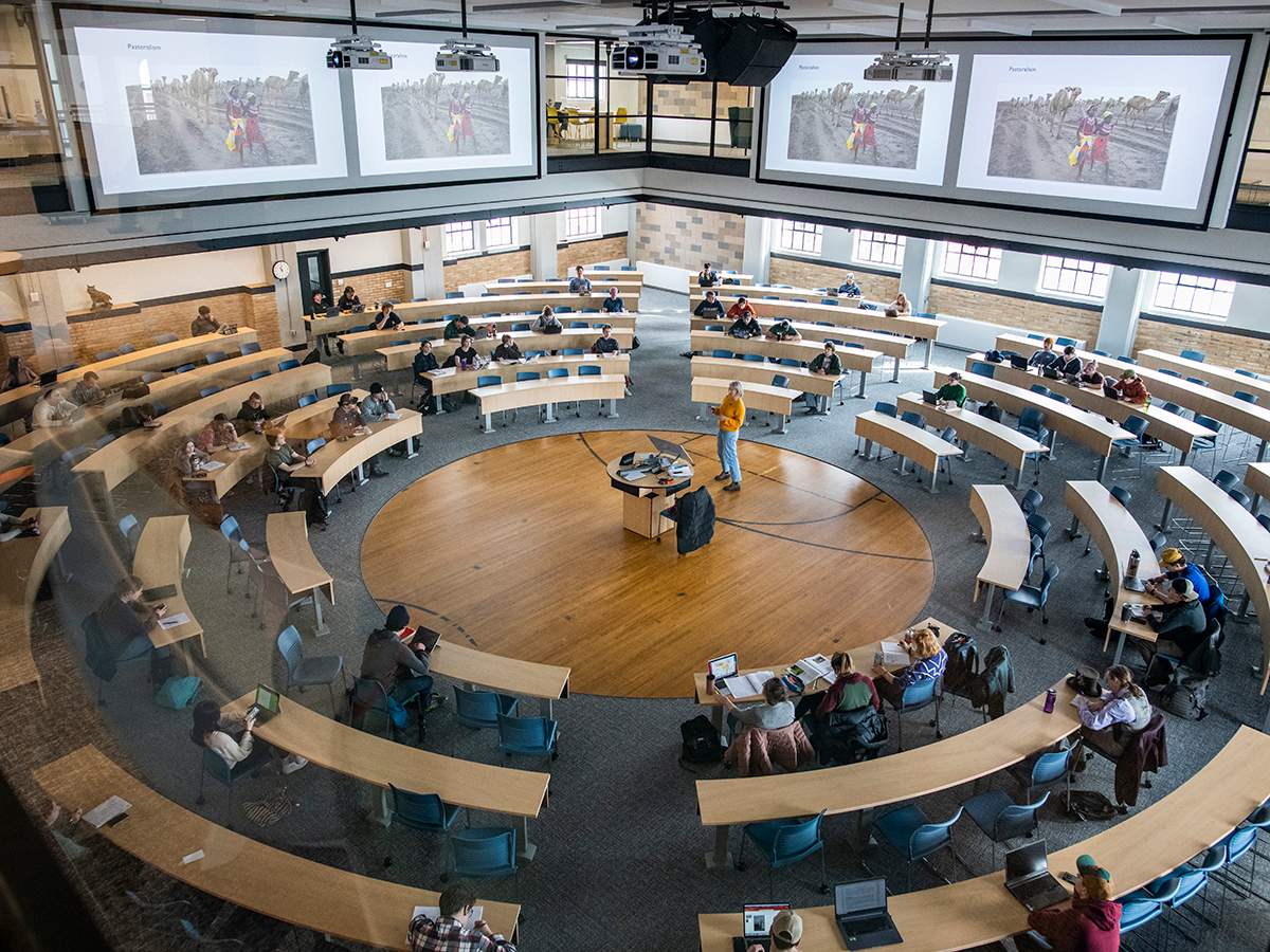 A circular classroom with a speaker in the middle. There are students at concentric tables taking notes.