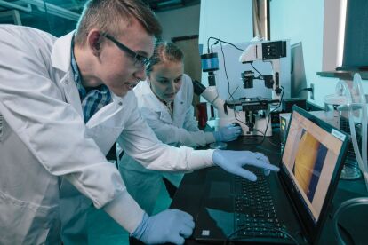 Two students in lab coats and gloves analyze data on a laptop next to a microscope in a laboratory.