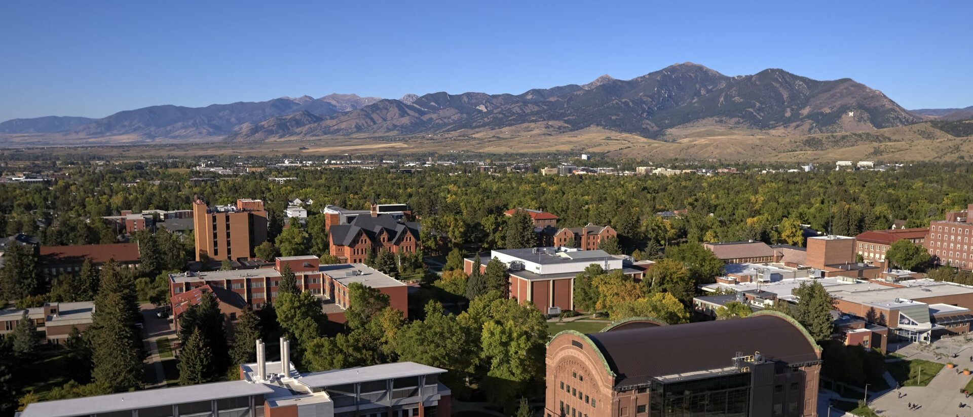 Aerial view of a university campus with multiple buildings and green trees, set against a backdrop of mountains und