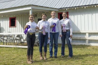 Four youth in white shirts and jeans stand outdoors holding ribbons and certificates at a county fair event.