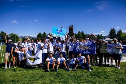 University sports team celebrates outdoors holding championship signs and flags under a clear blue sky.