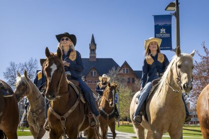 University equestrian team members ride horses on campus near a Montana State University banner and historic building.