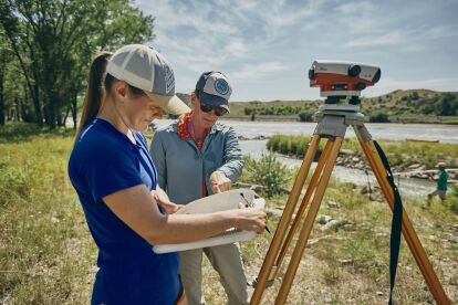 Two researchers record data near a river with surveying equipment set up on a tripod.