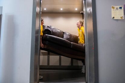 Two students in yellow shirts carry a large brown couch inside an elevator.
