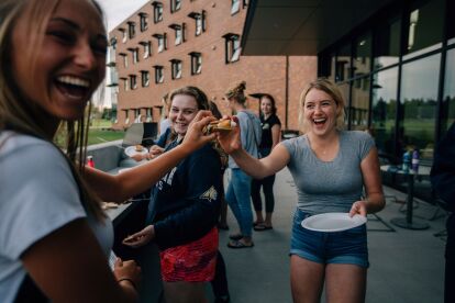 Students laugh and share food during an outdoor social event on a campus patio.
