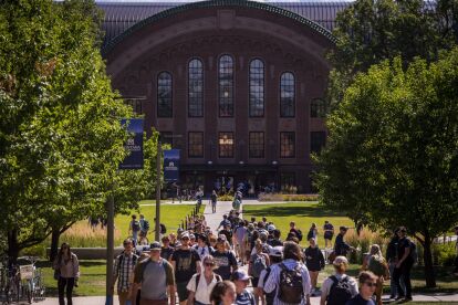 Students walk along a tree-lined path toward a large brick academic building on a sunny day.