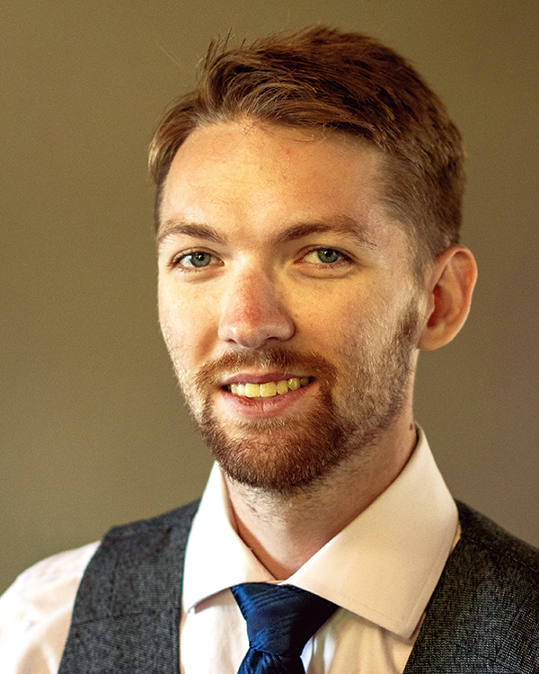 A person with short brown hair wearing a white collared shirt, a black suit vest, and a blue tie